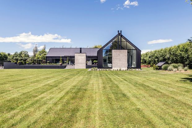 Wide exterior view of a contemporary low-profile home set behind an expansive manicured lawn. The building combines dark metal pitched roofing, charcoal cladding, large glazed openings, and prominent raw concrete wall volumes. A steep glazed gable forms a dramatic central architectural feature, with landscaped garden elements surrounding the house.