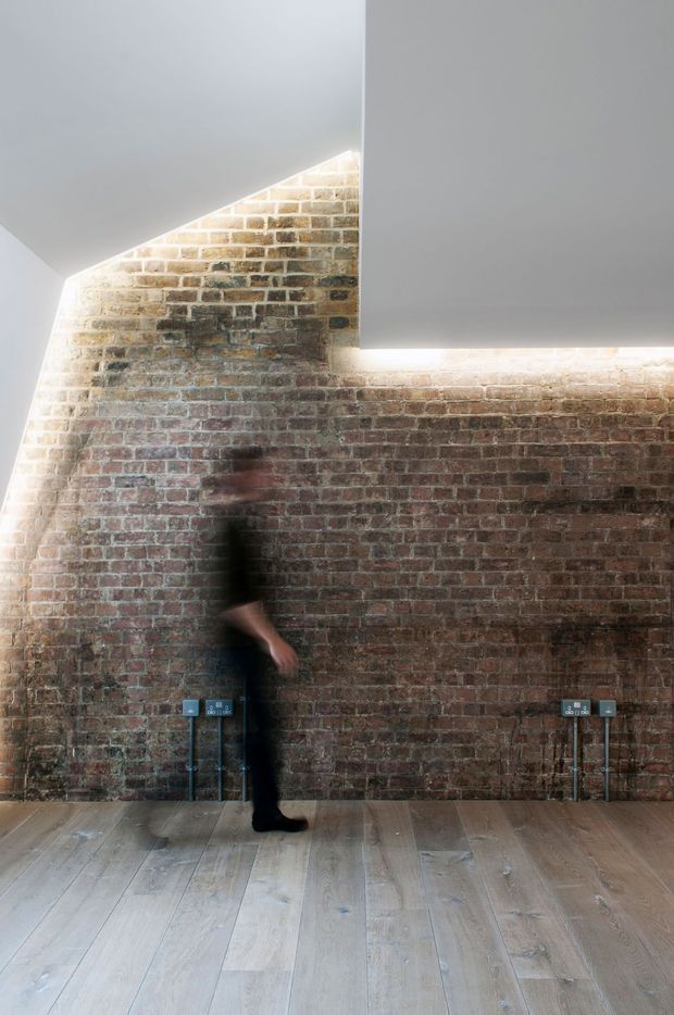 A minimalist interior with exposed aged brick walls, pale timber plank flooring, and crisp white angular ceiling planes. Linear concealed lighting washes the brickwork, while visible electrical outlets and a blurred person provide scale. The space has an industrial architectural character with a clean contemporary fit-out.