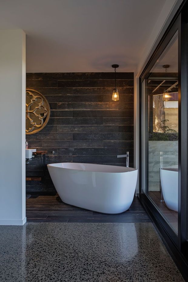 A moody contemporary bathroom featuring a freestanding white bathtub against a dark horizontal timber feature wall. The room has dark flooring, polished exposed-aggregate concrete in the foreground, black-framed full-height glazing with reflections, a wall-mounted tap, pendant lighting, and a decorative round wall feature.
