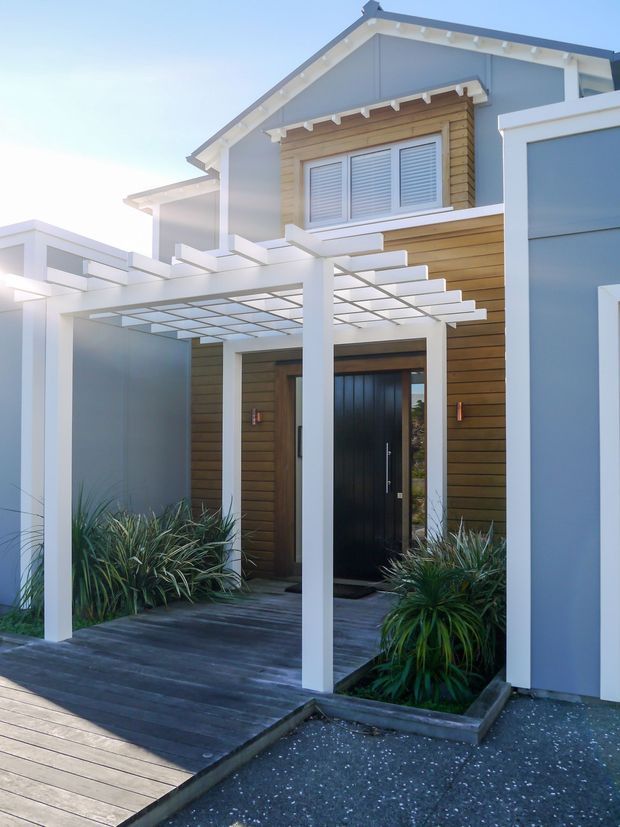 Contemporary two-storey residential facade with a gabled upper level, pale blue-grey cladding, warm horizontal timber weatherboards, white trim, and a black entry door. A white-framed pergola with translucent roofing extends over a dark timber deck and landscaped entry garden.