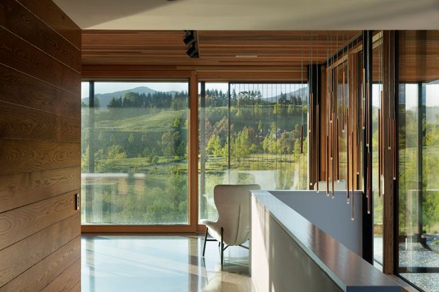 A contemporary interior with expansive floor-to-ceiling glazing overlooking a green, mountainous landscape. Warm timber wall cladding and a slatted timber ceiling frame a polished light floor, while a long dark counter or island occupies the right side. Vertical timber screens and suspended linear pendant lights add rhythm and privacy near the glazed edge. A light upholstered chair is positioned beside the counter.