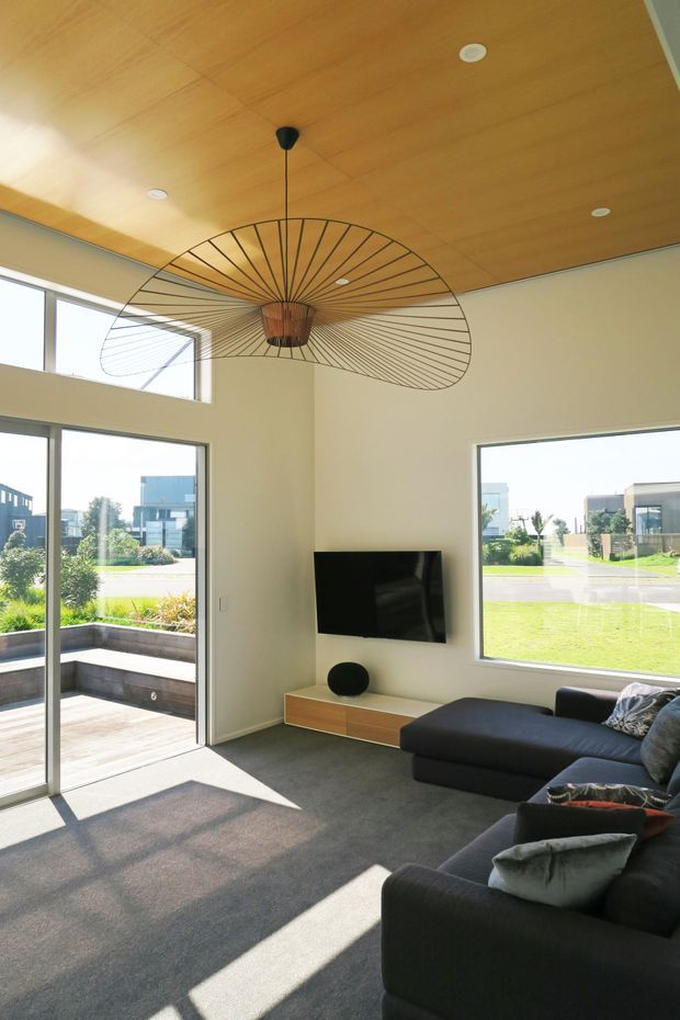 Bright contemporary living room with expansive corner glazing and a sliding glass door opening to a timber deck and landscaped exterior. The room features a warm timber-paneled ceiling with recessed downlights, pale walls, grey carpet, a large dark sectional sofa, wall-mounted television, low timber media console, and a striking oversized sculptural pendant light.