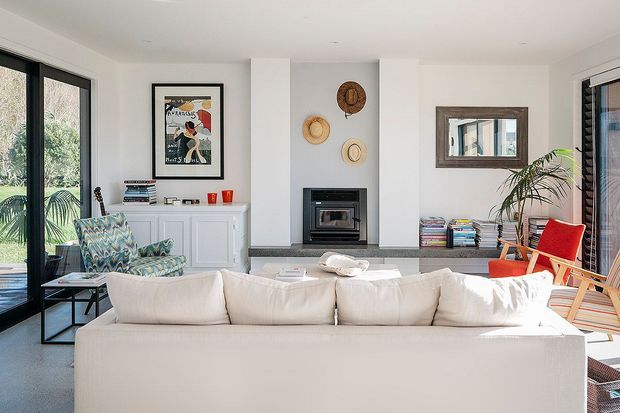 Bright, airy living room with a large cream sectional sofa in the foreground, patterned armchairs, a black-framed fireplace set into a white feature wall, built-in white cabinetry, expansive black-framed glazing, and a red accent lounge chair. Decorative wall pieces, books, and indoor plants add a relaxed contemporary character.