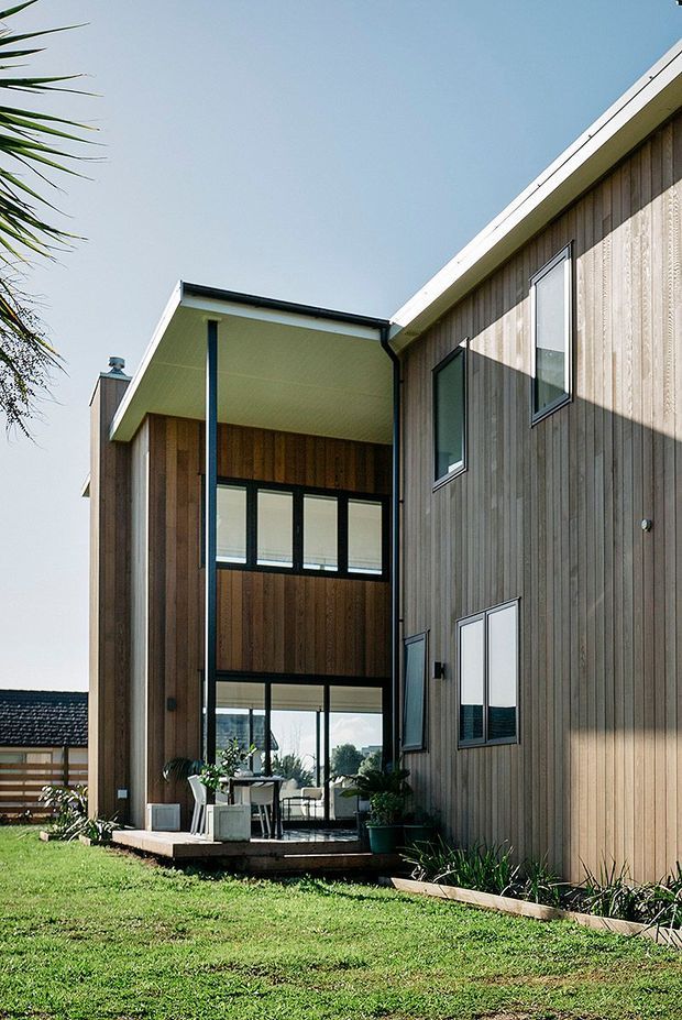 Exterior view of a contemporary two-storey residence with vertical timber cladding, a steeply angled roof form, black-framed glazing, and a recessed covered outdoor dining terrace. The home sits in a landscaped suburban setting with lawn and planting.