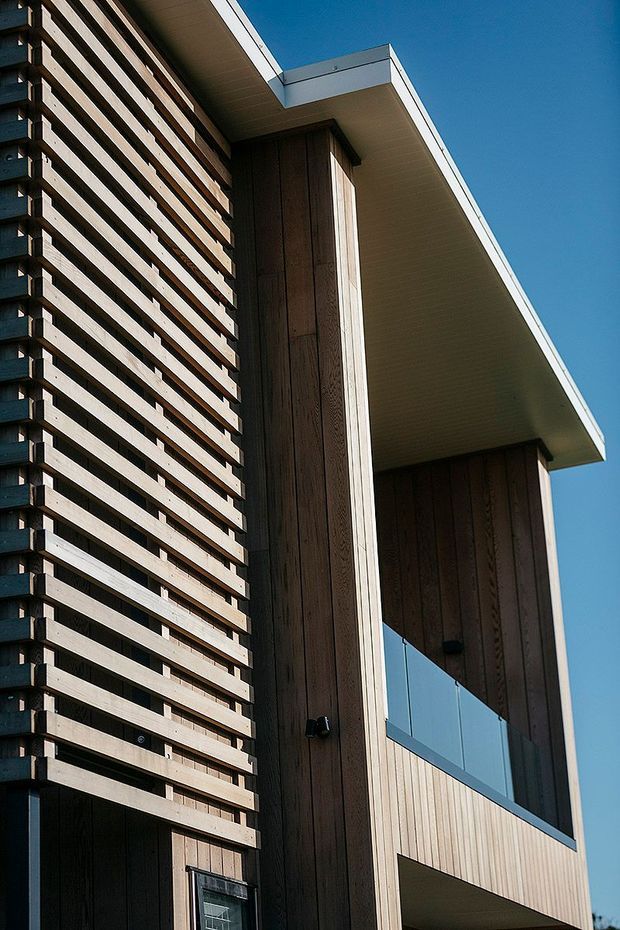 Close-up upward view of a contemporary building facade featuring vertical timber cladding, a projecting flat roof with a white soffit, horizontal timber screening, and a glass balcony balustrade against a clear blue sky.