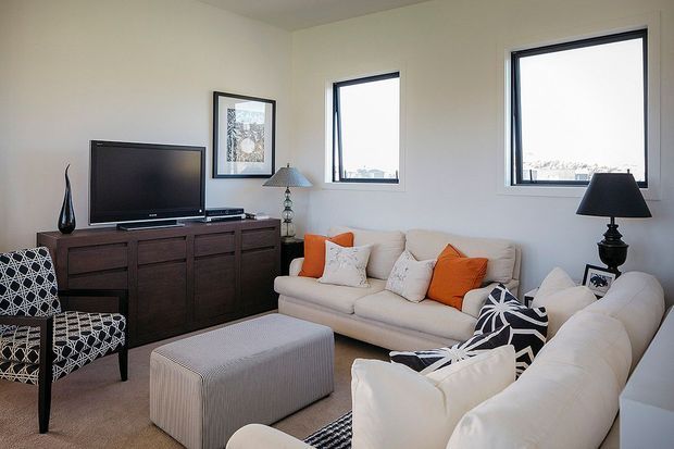 Bright contemporary living room with cream sofas, patterned black-and-white accent chairs and cushions, orange throw pillows, a textured ottoman, dark timber media cabinetry, framed artwork, table lamps, and three tall windows.