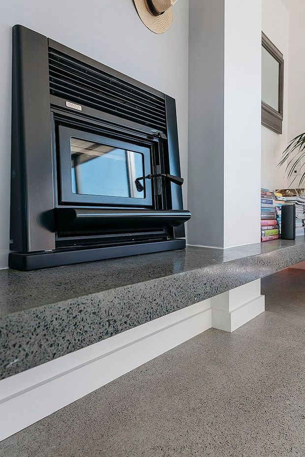 Close-up interior view of a black freestanding wood-burning stove set into a raised hearth or plinth. The stove sits on a thick grey speckled stone or terrazzo-like ledge, with white plaster walls, a polished grey floor, books, a framed artwork and a plant visible nearby. The space has a clean, contemporary residential feel with strong black, white and grey contrast.