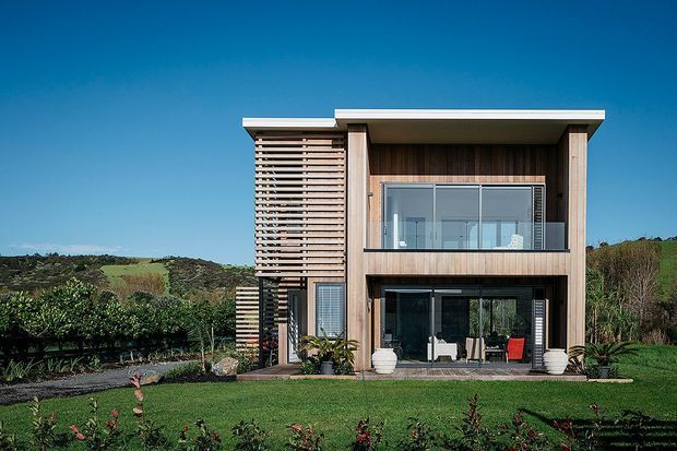 A modern two-storey residential house set within a landscaped garden, with a flat roof, extensive glazing, warm timber cladding, and a prominent horizontal timber screen on the upper level. The facade opens toward a lawn and planted garden, with rolling rural vegetation visible beyond.