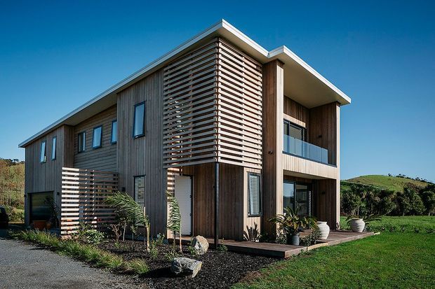 Modern two-storey residential home with dark vertical timber cladding, projecting flat roof forms, extensive horizontal timber screening, and a glazed upper-level balcony. The house is set in a landscaped rural or semi-rural site with lawn and low hills visible beyond.
