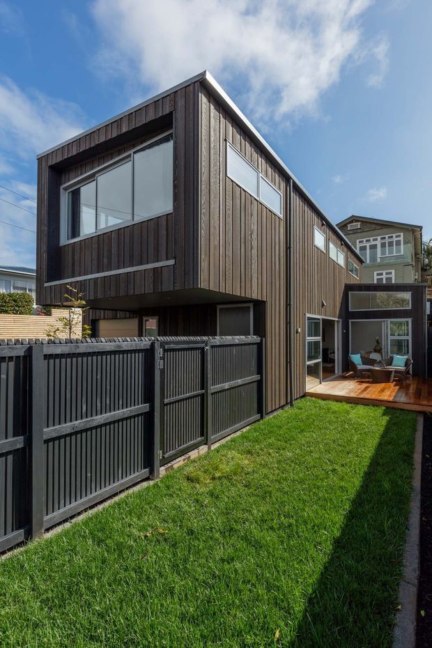 Exterior view of a contemporary multi-level residential extension or townhouse, clad in dark vertical timber boards with large glazed windows and doors. A dark slatted boundary fence runs along the grass verge, while a small timber deck and outdoor seating area are visible at the rear. The building has a simple pitched roof form and sits within a dense suburban setting under a bright blue sky.