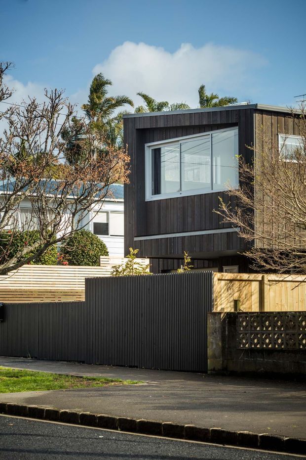 Street-facing view of a contemporary multi-level residential extension or townhouse, featuring dark vertical timber cladding, a projecting upper volume, large white-framed windows, and a contrasting pale timber boundary fence with black corrugated metal fencing. Mature suburban planting and neighbouring homes surround the property.