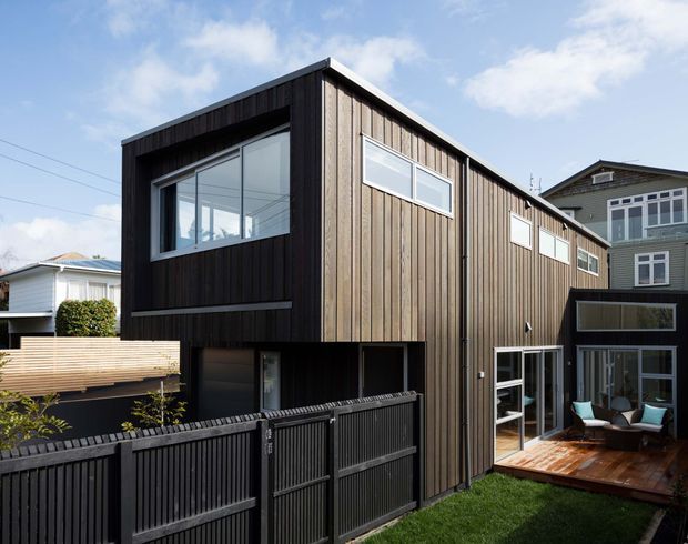 Exterior view of a contemporary multi-storey residential extension or townhouse with dark vertical timber cladding, large rectangular windows, a flat roof, and a small timber deck courtyard. A black timber fence and gate define the foreground, with neighbouring homes visible in the background.
