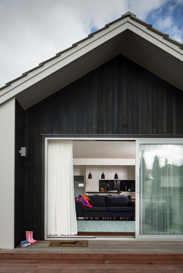 Exterior view of a gabled house with dark vertical timber cladding, white trim, and a broad glazed sliding door opening onto a timber deck. The open glazing reveals a contemporary living room with a dark sofa, rug, kitchen cabinetry, pendant lights, and built-in joinery.