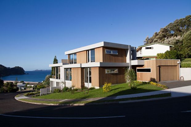 Exterior photograph of a contemporary multi-level residence on a sloping suburban coastal site. The home features strong horizontal roof planes, extensive glazing, warm vertical timber wall cladding, pale rendered bands, and an integrated timber-clad garage. A bay and wooded shoreline are visible in the background.
