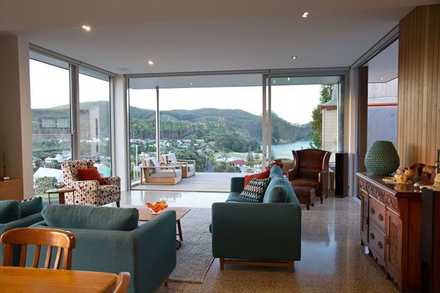 A spacious, light-filled living room with expansive floor-to-ceiling glazing overlooking a green, hilly landscape and water. The room features polished terrazzo flooring, teal upholstered sofas and armchairs, a patterned accent chair, timber dining furniture, a timber-paneled wall, and a traditional dark timber sideboard. Recessed ceiling lights and clean-lined glazing create a contemporary shell, while the furnishings add eclectic and mid-century influences.