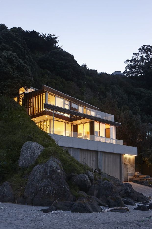 A contemporary multi-level house built into a steep, heavily wooded rocky site, photographed at dusk from a beach or rocky shoreline. The home features broad floor-to-ceiling glazing, warm interior lighting, projecting balconies and terraces, dark timber elements, pale concrete retaining walls, and an integrated lower-level garage. Large natural rock outcrops and dense vegetation surround the structure.