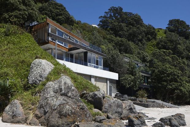Exterior view of a modern multi-level hillside house integrated into a steep, rocky, heavily vegetated site. The building features broad glazing, projecting balconies and decks, slender supports, horizontal concrete forms, and warm timber vertical screening beneath a shallow pitched roof. Large exposed rocks and a rocky shoreline or landscaped edge dominate the foreground.