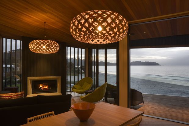 A warm, sophisticated living room overlooking the ocean through expansive floor-to-ceiling glazing. The room features a dark timber-lined ceiling and walls, a central fireplace with a lit fire, sculptural woven pendant lights, a dark sofa, yellow lounge chairs, and a timber dining table. A timber deck extends toward the beach and water beyond.