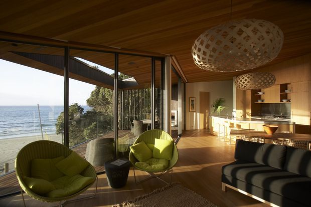A warm, open-plan coastal living and kitchen area with floor-to-ceiling glazing overlooking the ocean. The room features extensive timber wall and ceiling lining, timber flooring, a compact kitchen with island bench, woven pendant lights, lime-green lounge chairs, and a dark sofa. The architecture has strong indoor-outdoor connection and abundant natural light.