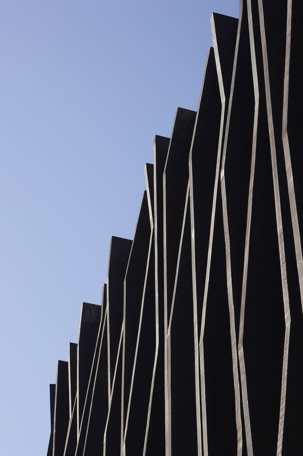 Upward-looking close-up of a striking dark, vertically articulated exterior facade. Repeating jagged or stepped panels create a strong geometric rhythm, with narrow light-coloured vertical strips contrasting against the near-black surfaces. Clear blue sky fills the background, emphasizing the building’s tall, sculptural form.