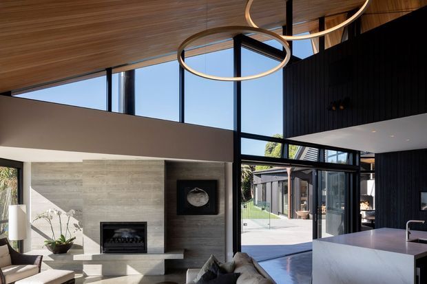 A sophisticated open-plan living space with a double-height volume, expansive black-framed glazing, and a warm timber-lined ceiling. The room includes a concrete or stone fireplace wall, contemporary fireplace, sculptural circular pendant lights, neutral furnishings, and a visible white kitchen island. An outdoor courtyard and additional building are visible through the glazing.