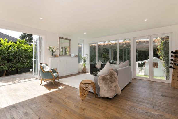A bright, spacious contemporary living room with extensive floor-to-ceiling glazing and open bi-fold doors connecting to a leafy outdoor area. The room features warm timber flooring, white walls and ceiling, a pale upholstered sofa with textured cushions and throw, a vintage-style armchair, woven side table, white sideboard, indoor plants, and integrated wine storage. Recessed ceiling lights and abundant daylight create an airy, inviting atmosphere.