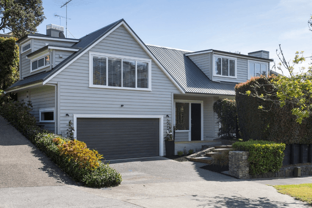 Street-level exterior view of a large light-grey weatherboard home with multiple gabled roof sections, a dark corrugated metal roof, white-trimmed windows, an integrated double garage, and a recessed entry. Neatly clipped hedges and low stone garden edging frame the driveway and front facade.
