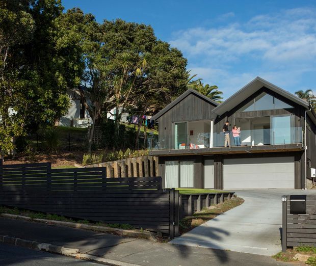 Exterior street-facing view of a contemporary multi-level home on a sloped suburban site. The house has dark vertical cladding, prominent gabled roof forms, large glazed openings, a balcony, and an integrated double garage. Tiered retaining walls, fencing, driveway, and established trees surround the property.