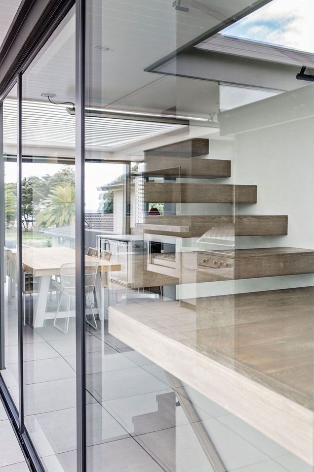 A bright contemporary kitchen viewed through full-height glazing, featuring a large timber-toned island or benchtop, integrated cabinetry, a built-in oven and cooktop, and distinctive floating timber shelves. Pale tiled flooring, white walls and ceiling, black-framed glazing, and an adjacent dining area create an open, refined interior with strong indoor-outdoor connection.