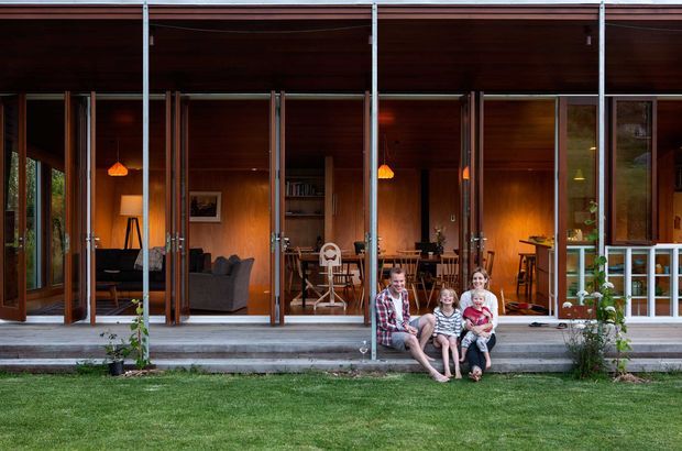 Exterior view of a contemporary timber-clad house with a deep covered veranda and extensive full-height glazed folding or sliding doors opening onto a lawn. Warm timber ceilings and walls, exposed slender structural posts, and an open-plan living and dining area are visible inside. A family sits on the broad concrete entry steps, with small garden plantings beside the facade.