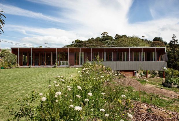 A long, single-storey contemporary house set within a landscaped garden. The building has a low-pitched roof, extensive floor-to-ceiling glazing and dark timber-framed openings, with a projecting enclosed section clad in grey metal or fibre-cement panels. A broad lawn, planted garden and mature vegetation surround the home.
