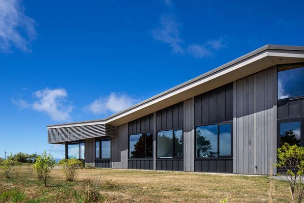 A low-profile contemporary building photographed from the exterior, featuring dark vertical cladding, expansive dark-framed glazing, a broad shallow roof plane, and a projecting louvred entrance canopy. The building sits in an open landscaped setting under a bright blue sky.
