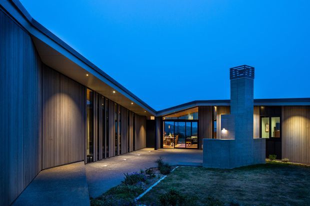 Twilight exterior view of a low-profile contemporary residence arranged around a sheltered courtyard. The building features dark vertical timber-like cladding, extensive floor-to-ceiling glazing, strong horizontal rooflines, recessed warm lighting, and a prominent rendered or masonry chimney volume. A central glazed entry connects the wings of the house, with sparse landscaping in the foreground.