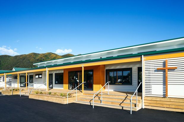 Single-storey contemporary building viewed from the exterior, with a long low-slung form, shallow roof, bold green and yellow trim, timber-clad entry sections, horizontal white cladding, large windows, and accessible ramped timber decking. A prominent timber cross feature is mounted on the right-hand facade. The building sits against a hilly landscape under a clear blue sky.