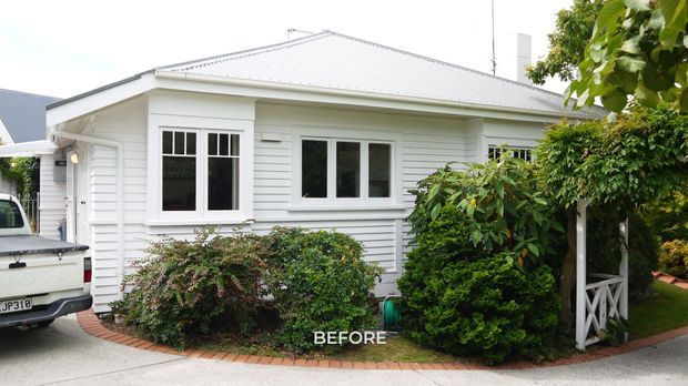 Exterior photograph of a modest single-storey white weatherboard house, likely a bungalow, with a low hipped metal roof, white-framed windows, covered side entry, and established shrubs along a curved brick-edged garden bed. A driveway and utility vehicle are visible in the foreground, and the image is marked “BEFORE.”