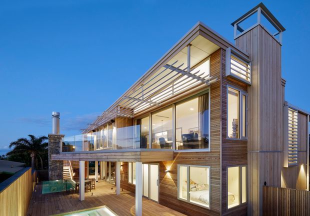 Contemporary multi-level residential house photographed from the exterior at dusk, featuring extensive floor-to-ceiling glazing, warm horizontal timber cladding, a large elevated terrace with glass balustrades and pergola-like screening, a tall vertical tower element, and a partially visible pool deck. The design has strong indoor-outdoor connections and dramatic architectural lighting.