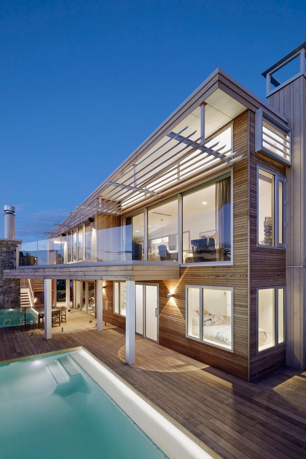 Exterior architectural photograph of a multi-level contemporary residence at dusk, clad in horizontal timber boards with extensive floor-to-ceiling glazing. A large timber deck, swimming pool, covered outdoor area, balcony, projecting upper volume, and pergola-like horizontal sun-shading structure are visible. Warm interior lighting contrasts with the deep blue evening sky.