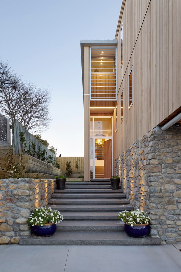 A contemporary multi-level home entrance approached by a broad flight of dark stone or concrete steps. The facade combines vertical timber cladding, extensive glazed entry doors and a projecting screened upper-level balcony or stair enclosure. Rough natural-stone retaining walls flank the stairs, with integrated step lighting and potted flowers adding warmth to the landscaped entry.