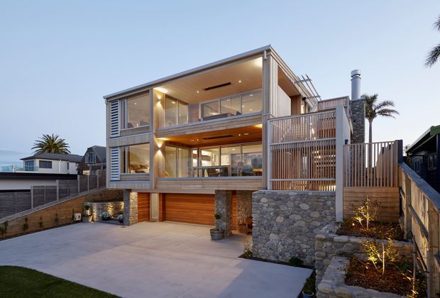 Exterior view of a contemporary multi-level residential house at dusk, featuring extensive glazing, warm timber soffits and screening, stone-clad structural elements and retaining walls, a large garage, balconies, and layered outdoor decks. The home sits on a sloping suburban site with a broad concrete driveway and landscaped planting.