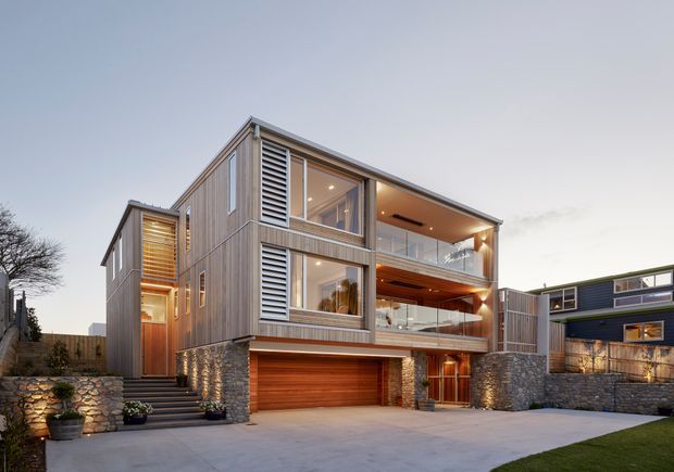 Contemporary multi-level residential house viewed from the front, featuring extensive glazing, vertical timber cladding, horizontal louvres, exposed warm timber soffits, stacked balconies, and a stone-clad base. A double garage and landscaped forecourt are prominent, with a neighbouring modern residence visible to the right.