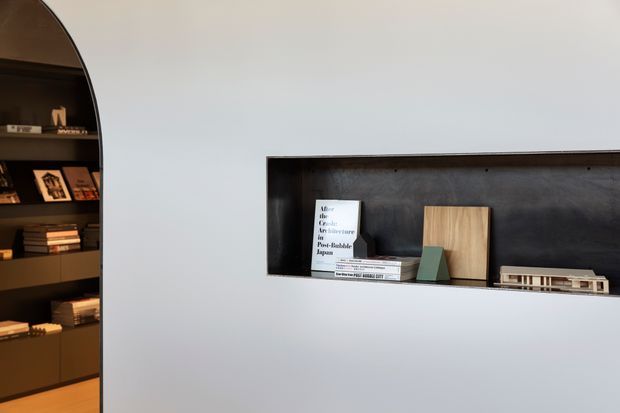 Minimal interior detail featuring a dark built-in arched shelving unit at the left and a long recessed wall niche on a pale wall. The shelving and niche display books, framed items, a small sculptural object, a timber panel, and architectural models. The composition is clean and restrained, with strong contrast between the dark joinery and light wall surface.