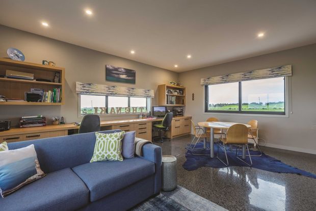A bright, open-plan multipurpose room combining a living area, home office, and dining zone. The room features a blue sectional sofa, built-in timber desks and shelving, a small round dining table with contemporary chairs, large windows overlooking a rural landscape, neutral walls, recessed ceiling lights, and a polished grey-speckled concrete or terrazzo floor.
