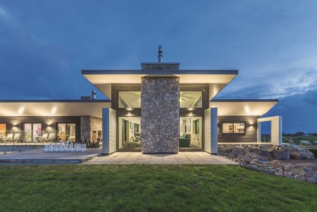 A contemporary single-storey house photographed at dusk, featuring a broad low-pitched roof with deep cantilevered eaves, extensive glazing, dark horizontal cladding, and a prominent central natural-stone-clad chimney or feature wall. Illuminated interiors and exterior soffits create a warm glow, with paved entry terraces and landscaped rock edging in the foreground.