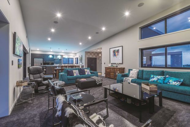 A spacious open-plan living room with high ceilings, large windows, dark carpet, teal sectional and sofas, a black leather lounge chair, glass coffee tables, timber sideboard, artwork, and a kitchen visible at the rear. Recessed lighting and contemporary furnishings create a polished, comfortable interior.