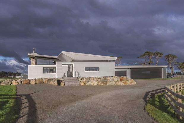 Single-storey contemporary residential home viewed from the driveway, with a light grey horizontal-clad facade, low pitched gabled roof, covered entry, dark integrated garage, stone retaining landscape, and expansive gravel forecourt. Dramatic overcast sky and rural-looking surroundings frame the building.