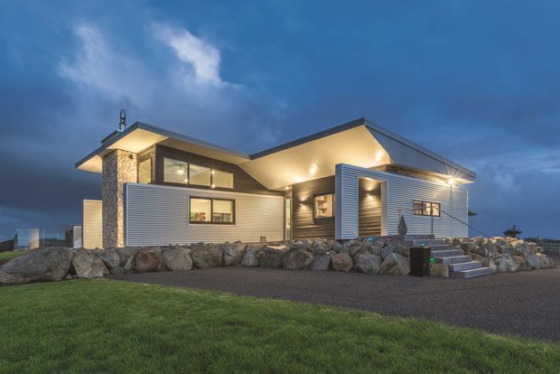 Exterior dusk photograph of a contemporary single-storey house with intersecting pitched roof forms, extensive horizontal metal cladding, a stone-clad entry tower, large glazed openings, and warm exterior lighting. The home sits behind a rocky retaining edge with a paved driveway and lawn in the foreground.