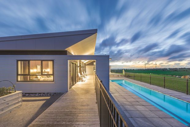 A contemporary single-storey building exterior at dusk, clad in dark horizontal metal siding with a sharply angled roof form and warm interior lighting visible through the windows. A timber walkway with black metal balustrades leads alongside a long rectangular swimming pool and paved pool deck, overlooking open rural fields.