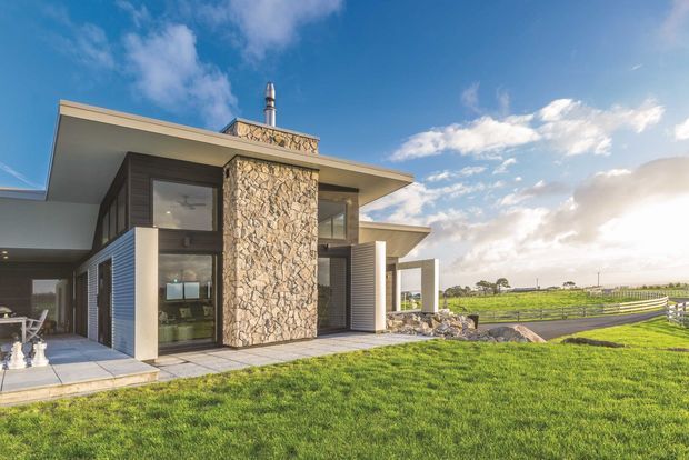 A contemporary single-storey house set in an open rural landscape. The facade combines broad flat roof planes, extensive glazing, horizontal grey cladding, pale rendered surfaces, and a prominent rough natural-stone chimney or feature wall. A covered outdoor terrace extends toward the lawn, with fenced fields visible beyond.