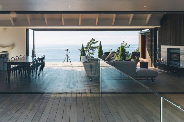 A refined open-plan living and dining area with full-width retractable glazing opening onto a terrace overlooking the sea. The room features dark timber flooring, a long dining table with black chairs, a charcoal sectional seating area, a glazed balustrade, exposed light timber ceiling beams, dark vertical timber wall panelling, and a built-in fireplace. Cypress-like trees and the ocean are visible beyond the terrace.