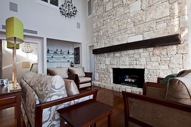 Formal living room with a large rustic light-stone fireplace wall, dark timber mantel and surround, polished dark timber flooring, and classic upholstered seating arranged around the fireplace. Built-in glass shelving displays bottles and glassware, while chandeliers and a tall green floor lamp add decorative accents. The room combines traditional architectural elements with refined, contemporary-classic furnishings.